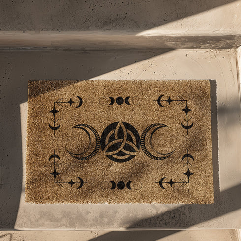 Rectangular coir doormat with central triple moon and triquetra symbol, surrounded by celestial motifs including stars and moon phases, in dark brown on natural tan background.